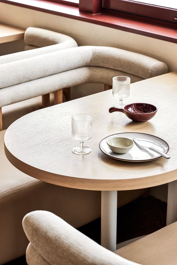 A warm, refined dining nook with a round light timber table, upholstered curved banquette seating and matching chairs, ribbed glassware, ceramic tableware, and a dark red serving bowl. A dark timber-framed window and soft beige upholstery complete the intimate setting.