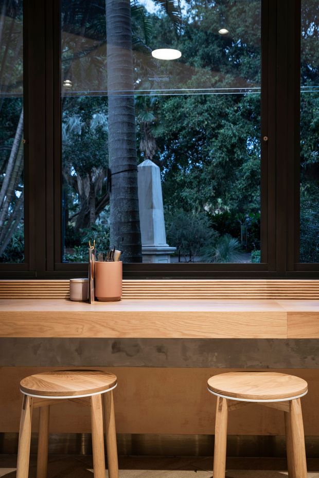 A compact, warmly lit interior workspace or casual breakfast nook set against a large dark-framed window overlooking dense greenery. A timber ledge and built-in bench run beneath the window, with two simple light-wood stools, a concrete-toned front panel, and a small container of stationery. Reflections of pendant lights are visible in the glazing.