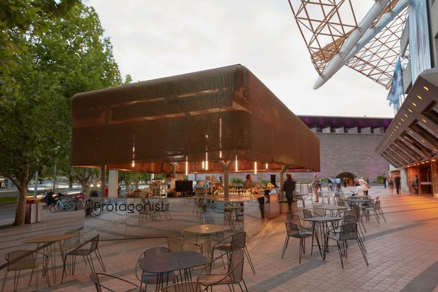 A contemporary outdoor dining and public plaza beside a commercial building. The open-air seating area is paved with large rectangular tiles and furnished with numerous black metal tables and chairs. A prominent suspended or elevated copper-toned perforated canopy encloses a bar or food-service area, with warm integrated lighting beneath it. The surrounding complex has modern angular structures, exposed steel roof elements, glazing, and mature trees at the edge of the plaza.