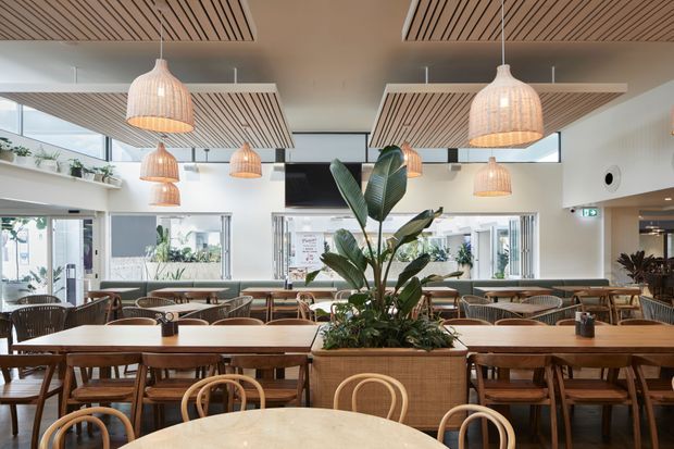 Bright contemporary commercial dining space with long communal timber tables, rounded timber chairs, woven pendant lights, indoor planting, and suspended timber slatted ceiling panels. Large windows and glazed openings create an airy, welcoming café or restaurant setting.