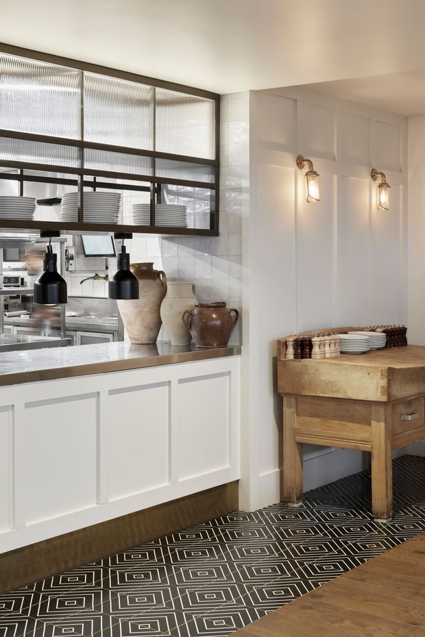 A refined restaurant or hospitality service area with white paneled cabinetry, a marble-topped counter, black industrial pendant lights, ribbed glass and dark metal-framed internal glazing, white tiled walls, and warm brass wall sconces. A rustic timber side table displays stacked plates and pottery, contrasting with the geometric black-and-white patterned floor and timber flooring.