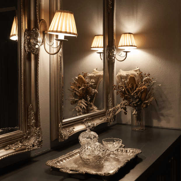 A warmly lit, ornate interior vignette featuring a dark console surface, large heavily embellished antique-style mirrors, decorative wall sconces with pleated fabric shades, and a silver serving tray with crystal glassware. A dried floral arrangement is reflected in the mirror, creating a luxurious, traditional atmosphere.