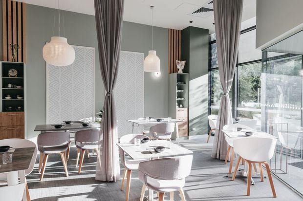 Bright contemporary dining room or restaurant interior with multiple neatly arranged dining tables and upholstered chairs. Floor-to-ceiling glazing brings in abundant natural light, while tall grey curtains divide the seating areas. The space features muted sage-grey walls, vertical timber screening, patterned decorative wall panels, built-in display shelving, pendant lights, and a light neutral fit-out.