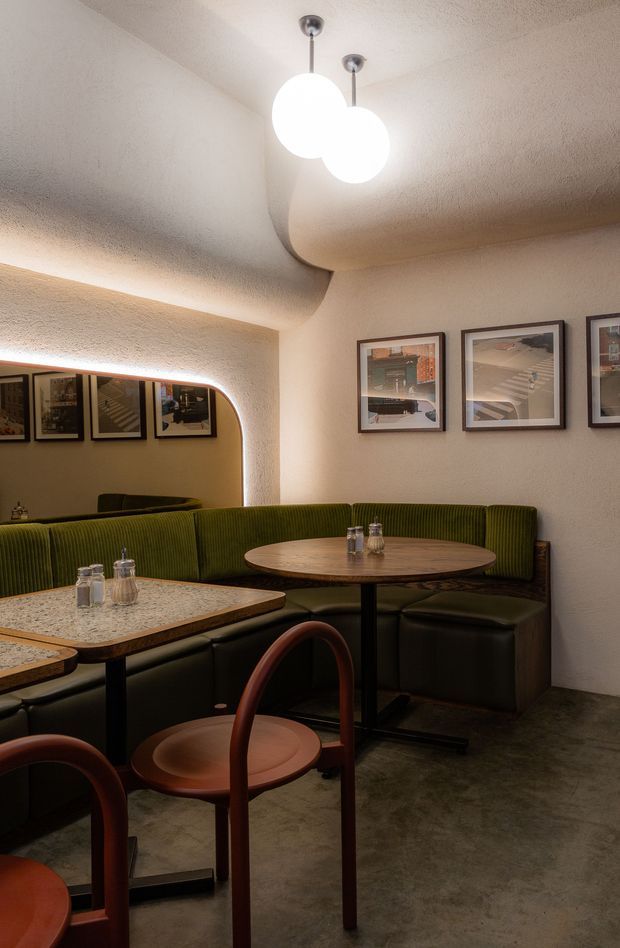 A warmly lit restaurant or café dining area with a curved green upholstered banquette wrapping around two small round tables. The space features textured cream walls and ceiling with sculptural curved detailing, a large rounded mirror with concealed lighting, framed artwork, polished stone tabletops, dark flooring, and suspended spherical pendant lights. Brown bentwood-style chairs occupy the foreground.