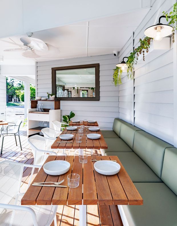 Bright casual dining area in a white-clad, partially open veranda or restaurant setting. Long timber dining tables are set with white bowls, plates, glasses and cutlery beside a built-in muted green upholstered bench. White metal chairs, hanging and wall-mounted greenery, ceiling fans, and a large dark timber-framed mirror contribute to a fresh coastal outdoor-dining atmosphere.
