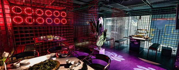 A dramatic, highly stylised open-plan dining and lounge interior enclosed by black metal grid screens. The space features vivid red and purple lighting, circular illuminated wall elements, industrial exposed ceiling services, large gridded windows, a dining table in the foreground, dark seating, a central decorative plant, and a compact bar or kitchenette area in the background.