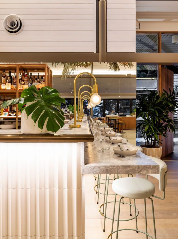A sophisticated restaurant or hospitality bar interior with a long marble bar counter, curved pale green metal stools, brass-toned arched pendant lights, ribbed white bar-front panelling, and extensive timber-framed glazing. A backlit liquor display and large tropical plant add warmth and a lush character to the refined space.