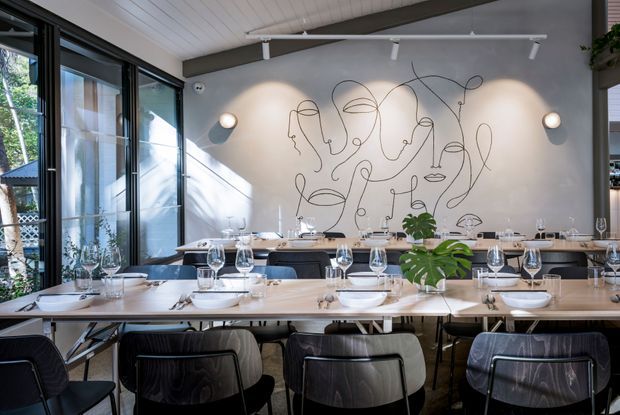Contemporary dining area in a restaurant or hospitality venue, featuring long communal timber tables set with dinnerware and wine glasses, dark upholstered dining chairs, a large expressive line-art mural on a grey wall, and extensive black-framed glazing along one side. A white-painted timber ceiling, exposed dark beam, track lighting, and indoor greenery complete the refined, modern setting.