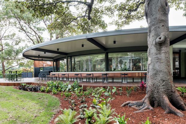 A modern single-storey pavilion-style building set within landscaped grounds, featuring a broad low-pitched roof with deep overhangs, dark structural framing, extensive glazing, and an outdoor covered terrace with a long bar counter and stools. Mature trees and planted garden beds surround the building.