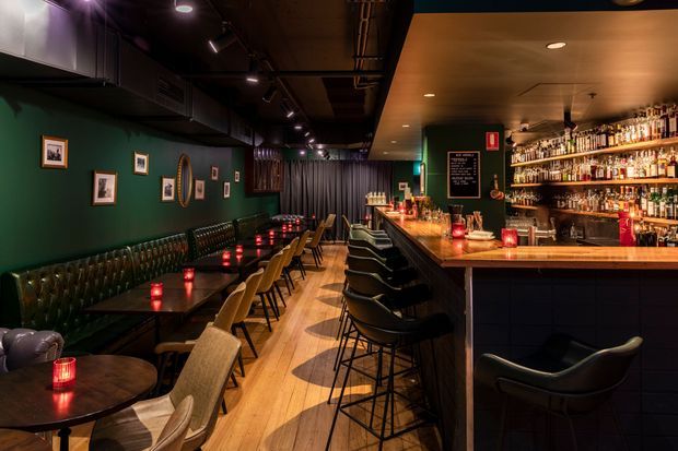 A dimly lit, intimate cocktail bar or restaurant interior with a long timber bar, extensive bottle display, black bar stools, dark green walls, tufted banquette seating, small round dining tables, framed artwork, and warm red candlelight. The space has an industrial ceiling with exposed services and track lighting, contrasted with polished timber flooring and a refined moody atmosphere.