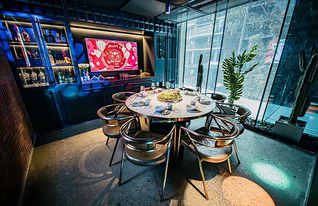 A dramatic contemporary dining area with a round set dining table and upholstered chairs, positioned beside floor-to-ceiling glazing. Dark built-in display cabinetry with illuminated shelving and a wall-mounted screen forms a feature wall, while colourful blue and warm accent lighting reflects across the polished concrete floor. Indoor planting and extensive glazing add contrast to the dark, atmospheric fit-out.