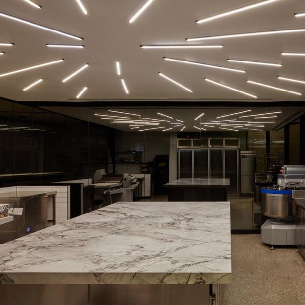 A dark, sophisticated commercial kitchen or food-service preparation space dominated by a large white-veined stone island bench. Stainless-steel appliances and preparation equipment line the perimeter, while black-framed glazing and mirrored surfaces reflect a dramatic ceiling installation of linear LED lights. The space has a refined industrial-contemporary character with a polished, high-end hospitality fit-out.