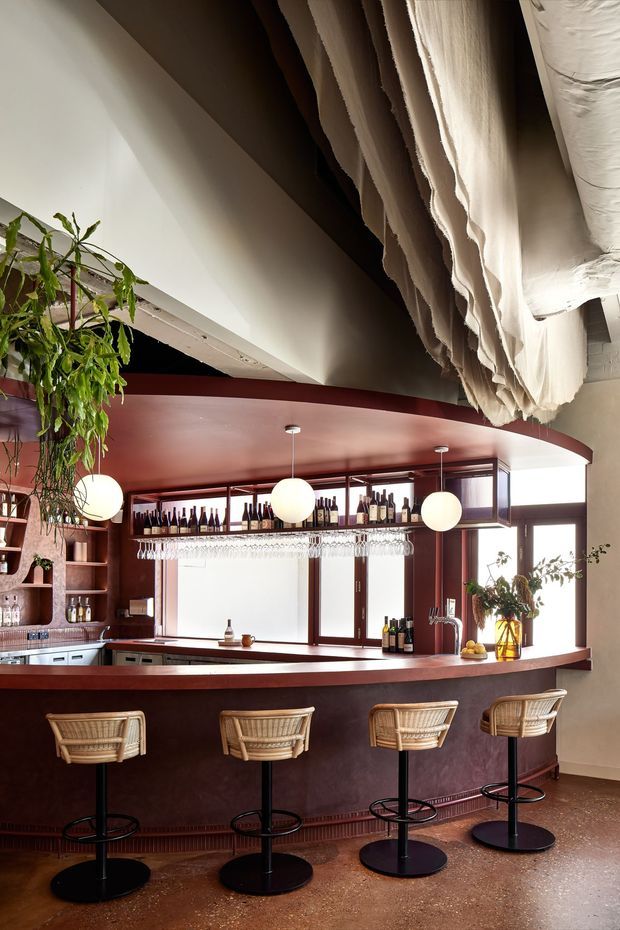 A stylish indoor bar area with a long curved burgundy bar counter, four woven rattan bar stools, built-in bottle shelving, hanging glassware, pendant globe lights, and large timber-framed windows. The space features a dramatic sculptural draped ceiling treatment, warm lighting, and polished terrazzo-like flooring.