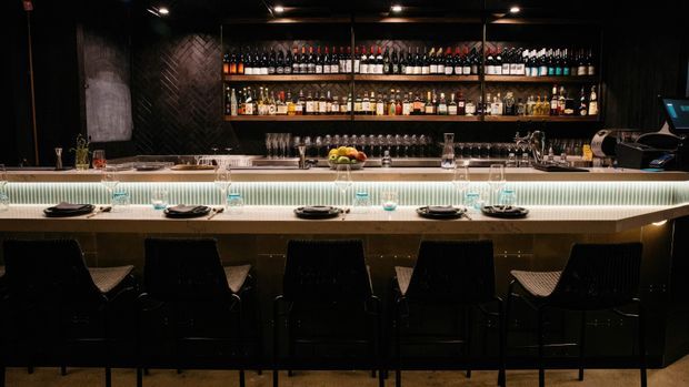 A dark, sophisticated bar and dining counter with a long light-coloured stone or composite benchtop, integrated linear lighting, black bar stools, and extensive open shelving displaying wine, spirits, and glassware. The space has a moody hospitality-inspired atmosphere with dark textured walls and black metal shelving.