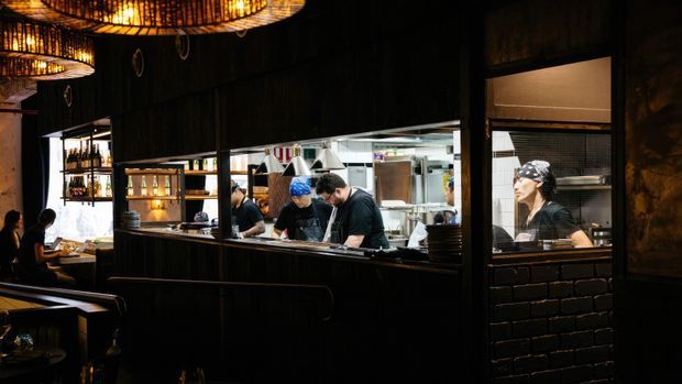 A dark, atmospheric restaurant interior showing an open kitchen behind a service counter, with chefs preparing food. The dining area includes booths and tables, while warm woven pendant lights, dark timber or stained wall surfaces, and black brick detailing create a moody hospitality setting.