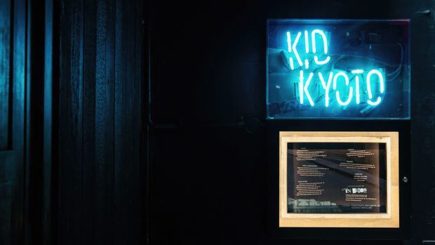 A dark, moody wall display featuring a bright cyan neon sign reading “KID KYOTO” above a framed poster or menu. The surrounding wall has black vertical timber-like panelling, creating a dramatic hospitality or entertainment venue atmosphere.