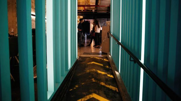 A narrow industrial-style interior passage or ramp lined with corrugated teal metal walls and vertical panels. A black handrail runs along the right side, while the dark floor features worn yellow directional chevrons. The passage opens into a dimmer social or commercial space with exposed timber and metal ceiling elements and several people in the distance.