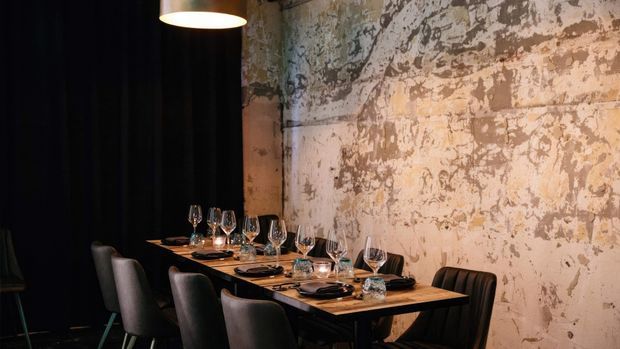 A dimly lit, intimate dining space with a long set table against a heavily weathered, textured plaster or concrete wall. Dark upholstered dining chairs, black curtains, warm pendant lighting, wine glasses, tableware, and candlelight create a refined industrial restaurant atmosphere.
