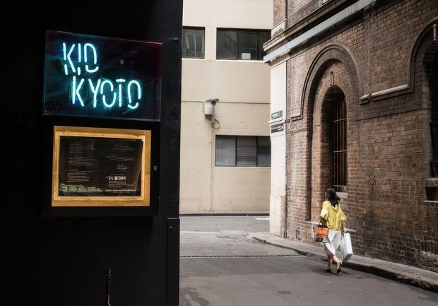 A narrow urban laneway beside a dark restaurant frontage displaying a blue neon “Kyo Kyo” sign and framed menu. A pedestrian walks past a historic brown-brick building with tall arched openings, while a plain contemporary building forms the background.