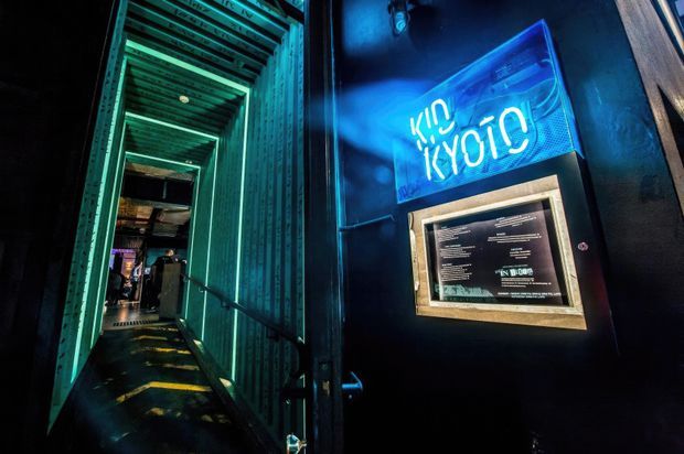 A dark, atmospheric entrance to a commercial venue, featuring a narrow passage framed by corrugated metal panels and illuminated with vivid turquoise and green strip lighting. A bright blue neon sign and illuminated menu or information display are mounted on the dark wall beside the entry.
