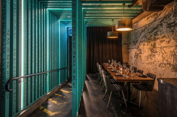 A dramatic commercial dining interior viewed through a narrow teal passageway. The restaurant features a long set dining table with place settings and wine glasses, dark upholstered chairs, warm pendant lights, black curtains, a distressed textured concrete or plaster wall, and dark flooring. The corridor has teal structural framing and Japanese-style vertical writing graphics, creating a strong industrial, atmospheric character.