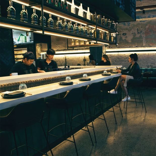 A dark, atmospheric contemporary bar and restaurant interior viewed along a long counter. The space features a pale stone or solid-surface bar top, dark upholstered bar stools, exposed textured masonry walls, open black metal shelving displaying bottles and glassware, and warm integrated shelf lighting. Staff work behind the counter while diners sit at the bar.