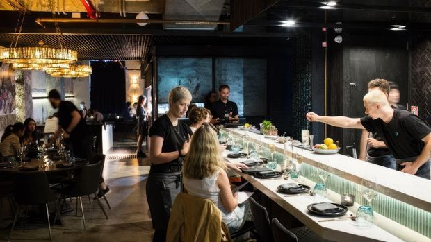 A dark, atmospheric restaurant interior centered on a long chef's counter with diners seated at the bar and staff preparing or serving food. The space features black walls and ceiling finishes, exposed services, dramatic pendant lighting, reflective surfaces, and an open-kitchen dining arrangement.