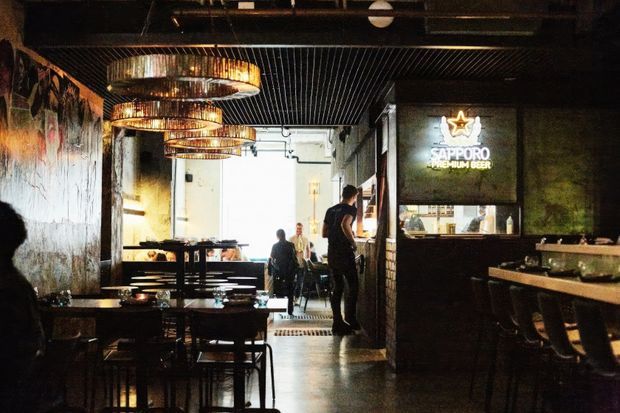 Dimly lit industrial-style restaurant interior with dark exposed ceiling services, black metal ceiling slats, textured feature walls, concrete-look flooring, pendant lights with woven or ribbed shades, timber tables, and a bar counter. A Sapporo beer sign is visible above the bar, with dining seating and patrons in the background.