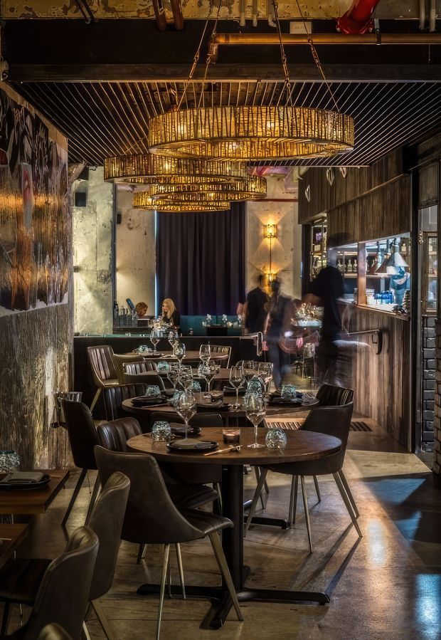 A moody, upscale restaurant interior with rows of round dark-wood dining tables, upholstered dining chairs, set tables with glassware, and a bar or service counter along the right wall. The space features dramatic tiered woven pendant lights, dark ribbed ceiling detailing, textured concrete and timber walls, warm ambient lighting, and a polished concrete floor. Diners and staff are present in the background.
