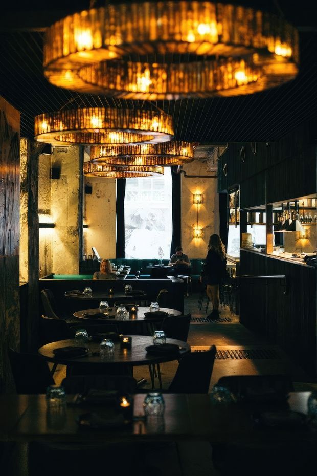 A dimly lit restaurant interior with rows of round dining tables and dark chairs, a long bar and service counter along the right side, textured plaster and dark wall finishes, and large woven circular pendant lights creating a warm amber glow. A projection screen and seated and standing patrons are visible in the background.
