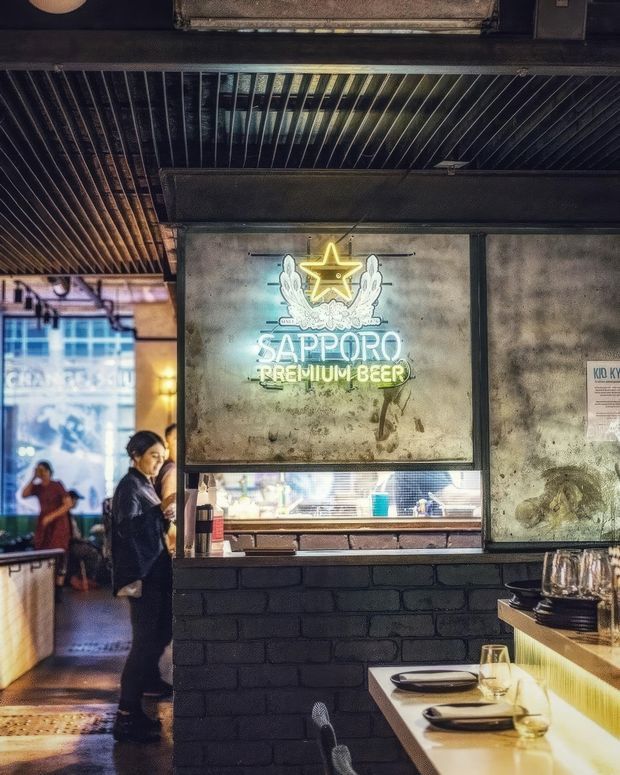 A dimly lit industrial-style restaurant or bar interior with a prominent illuminated Sapporo Premium Beer sign set into a distressed concrete service wall. A black-painted brick half-wall, dark ribbed metal ceiling, exposed ductwork, counter seating, plates, glasses, and a visible kitchen/service opening define the space. Diners and staff are present in the background.