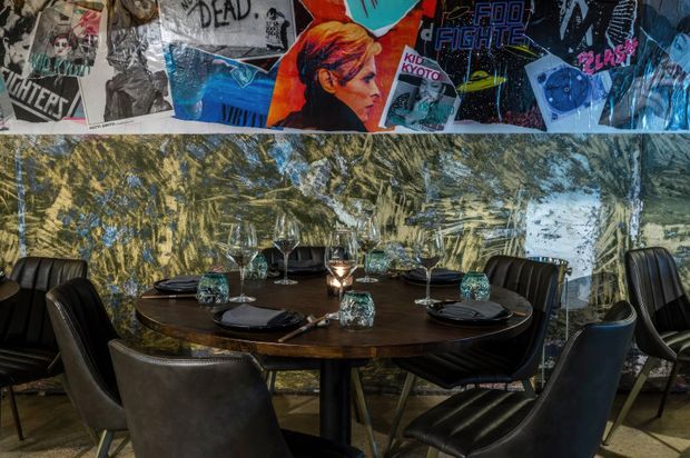 A moody dining area with a round dark timber table set for a meal, surrounded by black upholstered dining chairs. The space features a dramatic reflective marble-look feature wall and a colourful collage of graphic posters above it, with low candlelight creating an intimate atmosphere.