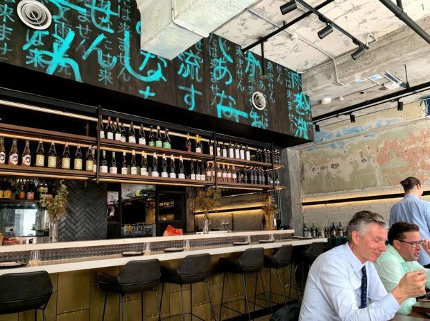A stylish bar and dining area with extensive bottle displays on open shelving, a long counter with high stools, and a textured industrial-style interior. The ceiling has exposed concrete and black track lighting, while the walls feature bold turquoise graffiti-like lettering and weathered mural finishes. Diners are seated at the bar and nearby tables.