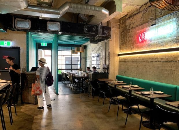 An industrial-style restaurant or café interior with exposed ductwork, rough painted brick and concrete surfaces, dark metal ventilation structures, and a polished concrete floor. Black dining tables and chairs line the room, with a long teal upholstered banquette beneath warm linear lighting and a neon wall sign. Large industrial-style windows and a glazed entrance bring light into the space.