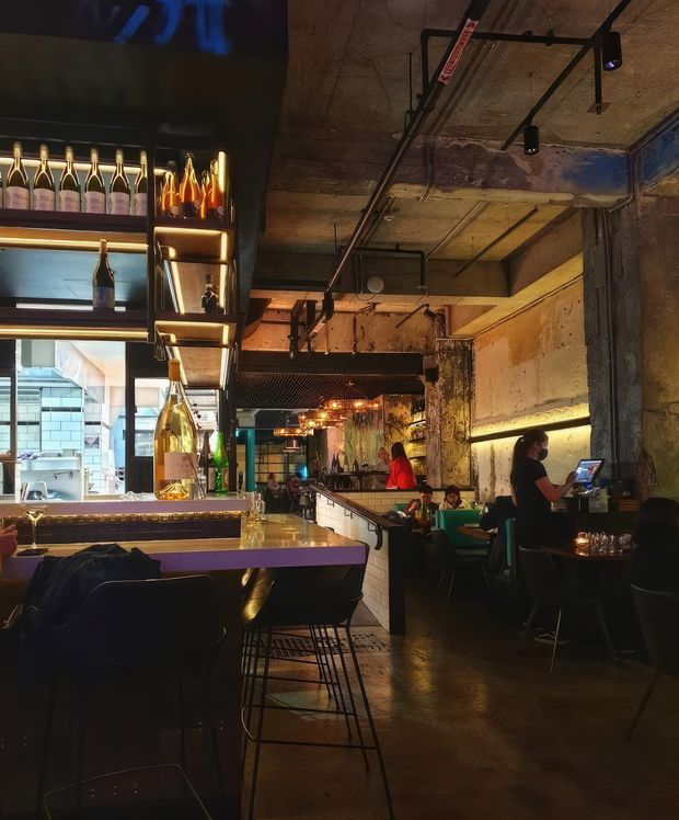 Interior view of a dimly lit, industrial-style bar or restaurant. The space features a long bar counter with high stools, illuminated bottle displays, exposed concrete and weathered plaster walls, exposed ceiling services, track lighting, booth seating, and warm pendant lights. Staff and patrons are visible, with a service counter extending through the centre of the venue.