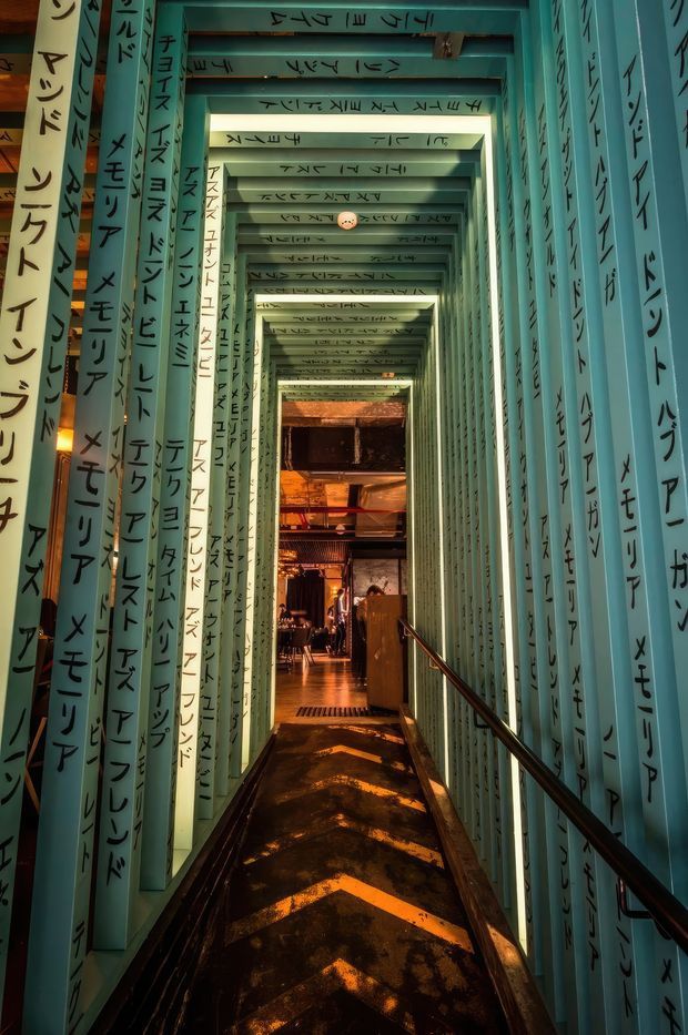 A narrow, immersive corridor or passageway lined with repeated turquoise structural frames covered in black handwritten or printed characters. The tunnel-like space recedes toward a warmly lit interior at the far end, with a dark floor marked by orange chevrons, metal handrails, and an industrial restaurant or bar area visible beyond. The scene has a dramatic, theatrical atmosphere with strong cyan-and-amber contrast.