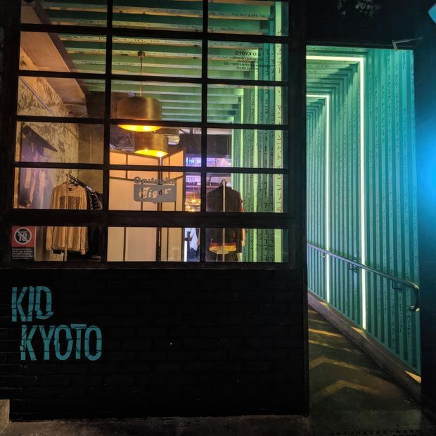 Night-time exterior view of a dark brick commercial storefront or venue, with large black-framed industrial grid windows revealing clothing racks, warm pendant lighting and exposed interior surfaces. A narrow illuminated passage with corrugated green wall panels and a handrail runs beside the building. Turquoise painted lettering appears on the brick facade.