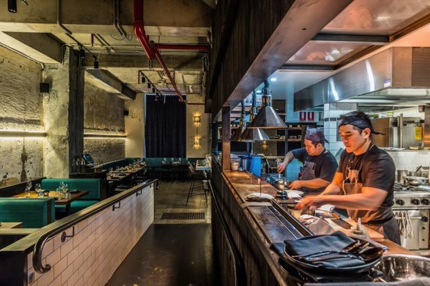 A dramatic industrial-style restaurant interior viewed from the dining area toward an open commercial kitchen. The foreground features tiled dining counters and dark green banquette seating, while chefs work behind a long stainless-steel counter under exposed metal ventilation equipment. Concrete walls, exposed services, black steel elements, warm pendant lighting, and dark finishes create a moody hospitality atmosphere.