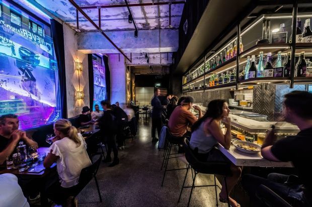 A busy, moody urban bar or restaurant interior with patrons seated at tables and along a long counter. The space features exposed concrete and structural elements, dark finishes, metal shelving stocked with bottles, patterned tile behind the bar, pendant and track lighting, and vivid blue-purple illuminated wall graphics.