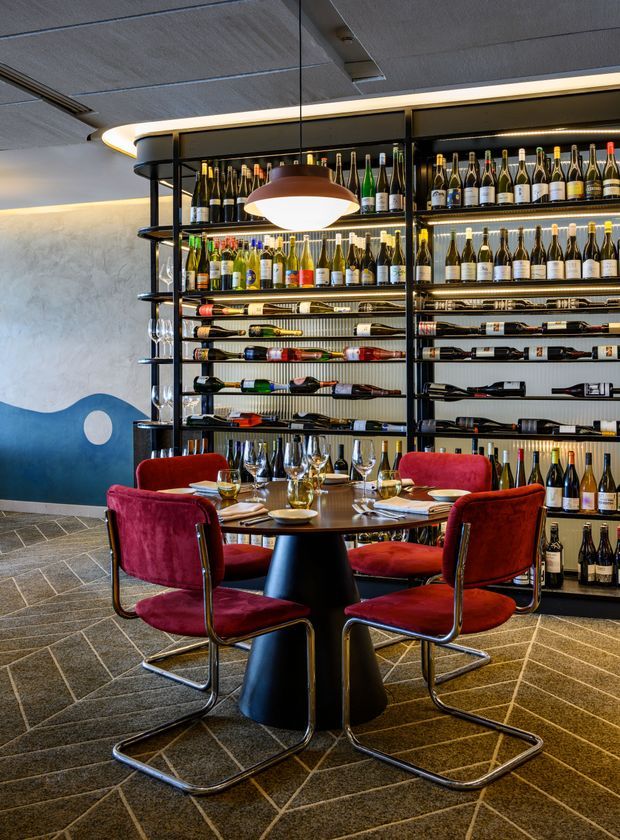 A stylish wine bar or restaurant dining area with extensive illuminated wine shelving covering the back wall. A round table is set with wine glasses and tableware, surrounded by plush red velvet cantilever chairs. The space features dark metal shelving, warm integrated lighting, a sculptural pendant light, textured wall finishes, and patterned dark flooring.