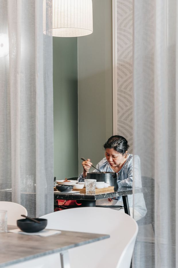 A softly lit dining area viewed through sheer curtains, with a round white dining table and chairs in the foreground. A person is seated at a smaller table behind it, eating from a bowl. The space features a muted green wall, patterned wallpaper panel, pale curtains, and a large cylindrical pendant light, creating a calm contemporary interior.