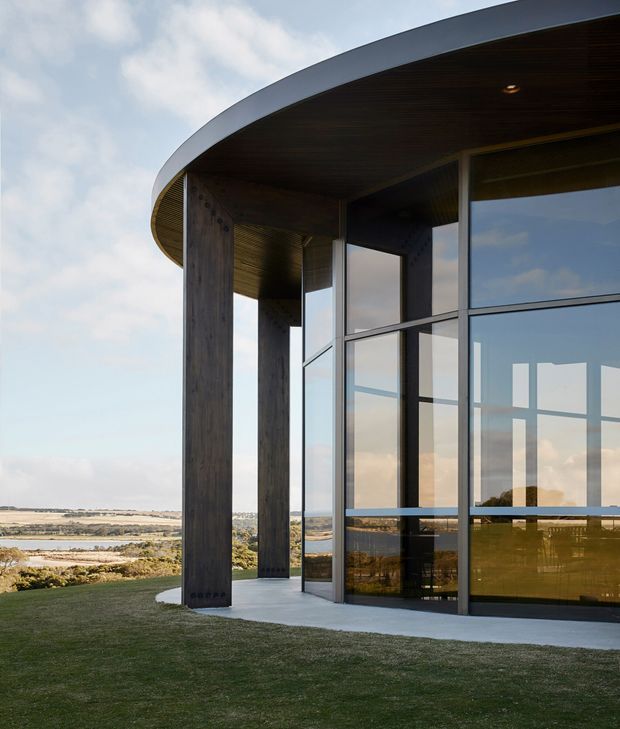 Exterior architectural photograph of a contemporary curved-roof building beside a broad coastal or lakeside landscape. The structure features large floor-to-ceiling glazed walls with dark aluminium framing, substantial dark timber or wood-clad columns, a deep timber-lined soffit, and a pale concrete base. The sweeping roof overhang and strong vertical supports create a sculptural, sheltered facade opening toward the landscape.