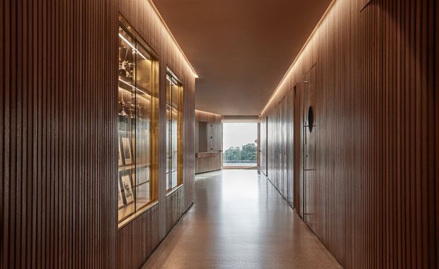 A long, narrow, warmly lit residential hallway lined with dark timber battens and integrated display cabinetry. Glass-fronted illuminated shelving is visible on the left, while flush timber doors and wall panels run along the right. The polished floor leads toward a bright glazed opening with greenery beyond, creating a refined, gallery-like corridor.