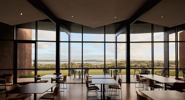 A contemporary café or dining space with extensive floor-to-ceiling glazing overlooking a coastal landscape. The interior features multiple light-coloured rectangular tables, dark-framed chairs, black structural columns, a dark ribbed ceiling, polished hard flooring, and warm recessed lighting. The expansive windows create a strong connection to the outdoor setting.