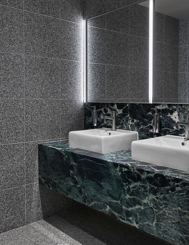 A compact contemporary bathroom featuring twin white vessel basins on a dramatic dark green veined stone vanity. The walls and floor are clad in dark grey speckled large-format tiles, while mirrored cabinetry and vertical integrated lighting create a polished, moody atmosphere.
