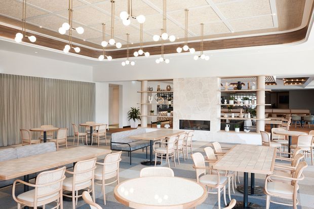 Bright, refined hospitality-style dining and lounge interior with numerous light timber tables and woven-seat chairs arranged around a central stone-clad fireplace and open shelving. A curved timber ceiling detail with suspended globe lights frames the space, while full-height curtains, pale upholstery, and terrazzo-like flooring create an airy, warm atmosphere. A bar or service area is visible at the rear.