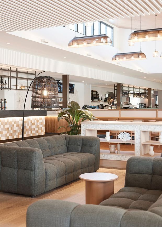 Bright, spacious hospitality lounge and bar interior with a double-height ceiling, extensive timber slatted ceiling detail, high clerestory glazing, and multiple suspended linear pendant lights. The foreground contains deep grey upholstered lounge seating and a small round timber coffee table. Behind it is a refined bar and service counter with warm timber finishes, light patterned tile detailing, open shelving, decorative objects, and indoor planting.