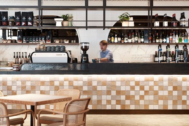 A stylish café or bar interior with a long service counter, espresso machine, coffee and liquor shelving, and a staff member behind the counter. The space features a black-framed glazed screen and shelving, a marble-look backsplash, a checkerboard mosaic-tile counter front in cream, beige and terracotta tones, and light wood café tables with woven rattan chairs.