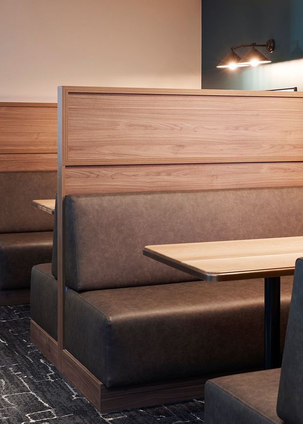 A refined booth-style commercial dining interior with built-in dark brown upholstered banquette seating, warm timber privacy screens and tables, dark patterned carpet, and a wall-mounted brass-toned light fixture. The composition focuses on the fixed booth joinery and seating arrangement.