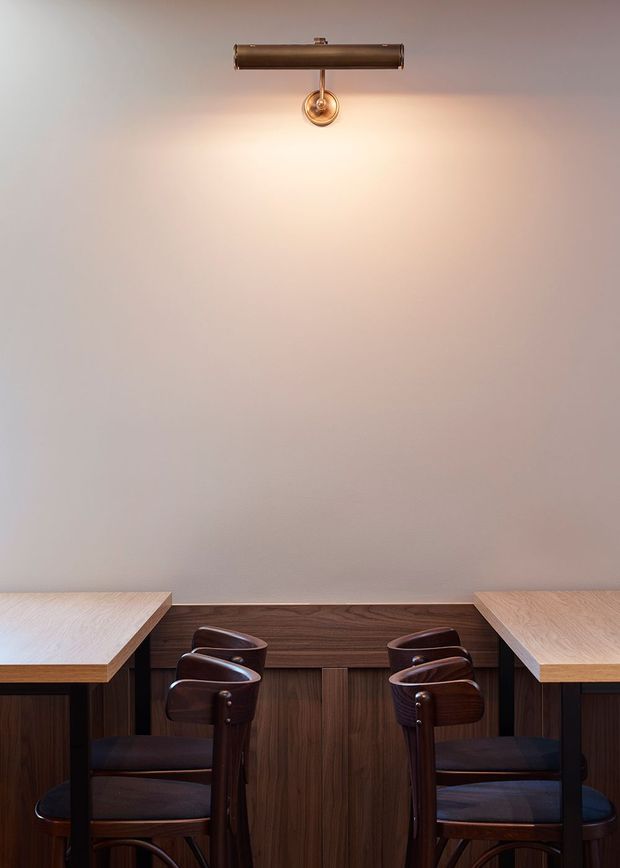 A compact, symmetrical dining or café-style interior with two light timber tables facing each other, dark wood cabinetry or panelled joinery along the back wall, and paired dark upholstered wooden chairs. A brass-toned wall light provides warm illumination against a plain pale plaster wall.